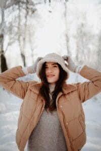 Woman in warm winter clothing, smiling amidst a snowy backdrop, showcasing a cozy and fashionable look.